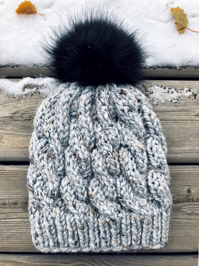 May include: A grey and white cable knit beanie with a black faux fur pom pom. The beanie is made from a soft, warm yarn and is ideal for cold weather.