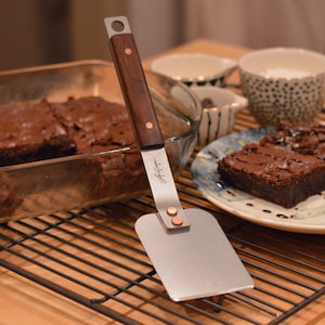 May include: A metal spatula with a wooden handle is used to lift a piece of chocolate brownie from a glass baking dish. The brownie is on a white plate with a brown and green design.