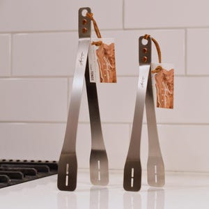 May include: Two stainless steel tongs with a leather loop and a tag. The tongs have a sleek, modern design with a small logo on the side. The tags have a brown and white design. The tongs are on a white surface.