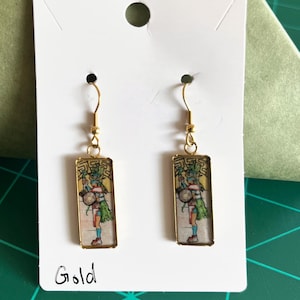 May include: Gold-colored rectangular earrings with a colorful illustration of a figure in traditional clothing. The earrings are displayed on a white card with the word "Gold" written on it.