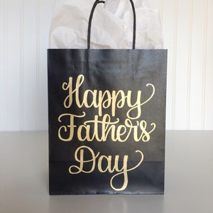 May include: Black gift bag with black handles and white tissue paper inside. The bag has gold lettering that says "Happy Father's Day."
