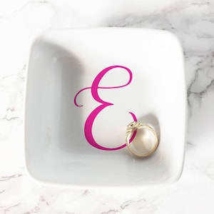 Könnte beinhalten: Eine weiße Keramik-Ring Schale mit einem pinkfarbenen, kursiv geschriebenen Buchstaben "E" und einem goldenen Ring im Inneren.