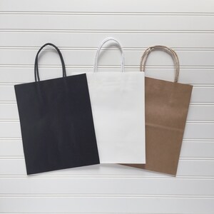 May include: Three paper shopping bags in black, white, and brown. Each bag has a rectangular shape with a matching handle. The bags are arranged against a white, horizontal-slatted background.