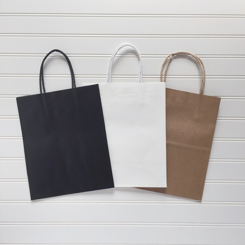 May include: Three paper shopping bags in black, white, and brown. Each bag has a rectangular shape with a matching handle. The bags are arranged against a white, horizontal-slatted background.