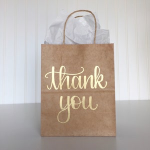 May include: A brown paper gift bag with a gold "thank you" inscription. The bag is filled with white tissue paper.