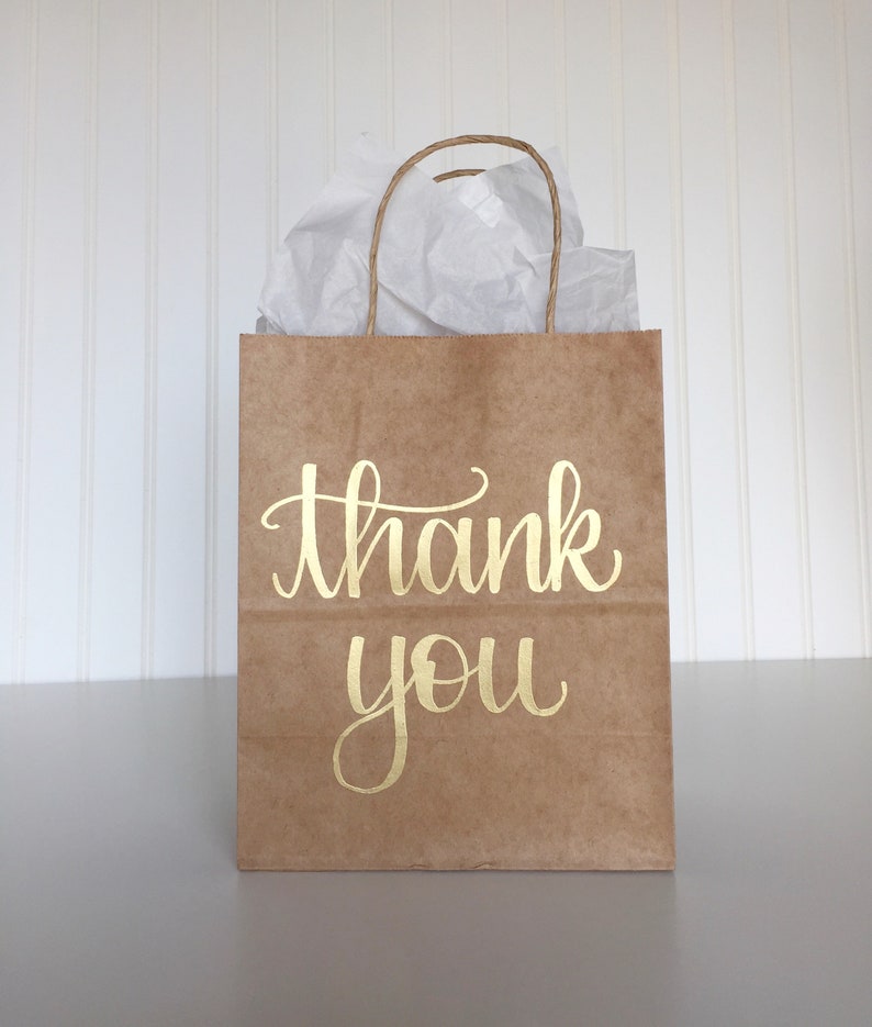 May include: A brown paper gift bag with a gold "thank you" inscription. The bag is filled with white tissue paper.
