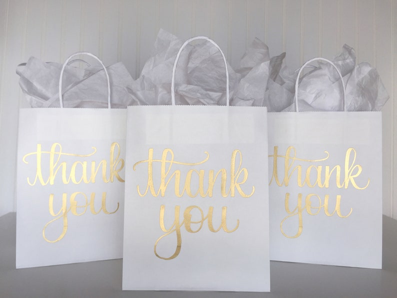 May include: Three white paper gift bags with gold foil lettering that says "thank you".