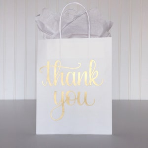 May include: White paper gift bag with gold foil lettering that says "thank you".