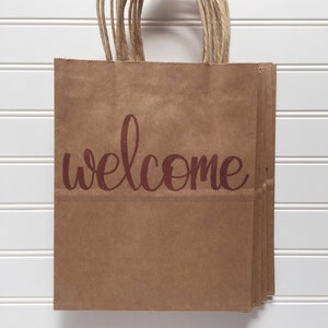 May include: A stack of brown paper gift bags with twisted paper handles. The word "welcome" is printed in a burgundy script across the front of the bags. The bags are displayed against a white, slatted background.