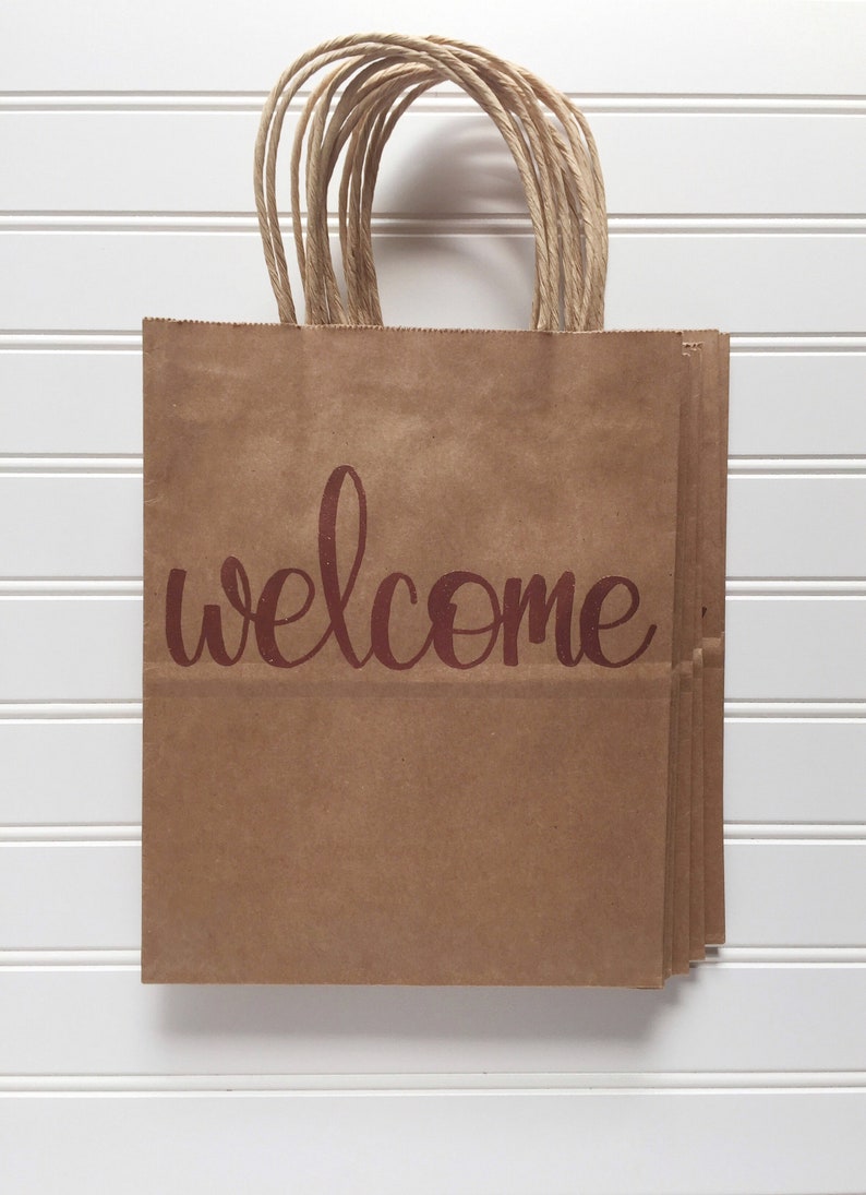 May include: A stack of brown paper gift bags with twisted paper handles. The word "welcome" is printed in a burgundy script across the front of the bags. The bags are displayed against a white, slatted background.