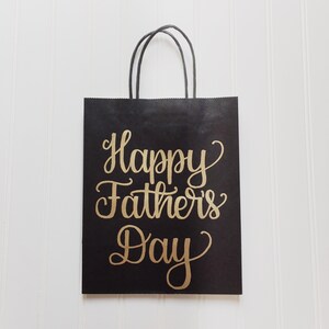 May include: Black paper gift bag with a gold inscription that reads "Happy Father's Day".