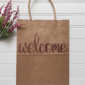 May include: A brown paper gift bag with twisted paper handles. The word "welcome" is printed in a copper-colored script across the front. A sprig of pink flowers is in the upper left corner, set against a white background.