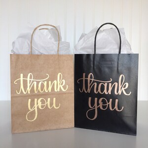 May include: Two gift bags, one brown and one black, with the words "thank you" written in gold and rose gold lettering.