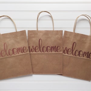 May include: Three brown paper gift bags with twisted paper handles. Each bag has the word "welcome" printed in a cursive font in a reddish-brown color. The bags are arranged on a white, slatted surface.