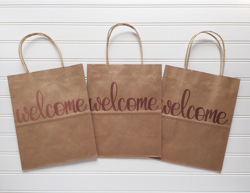 May include: Three brown paper gift bags with twisted paper handles. Each bag has the word "welcome" printed in a cursive font in a reddish-brown color. The bags are arranged on a white, slatted surface.