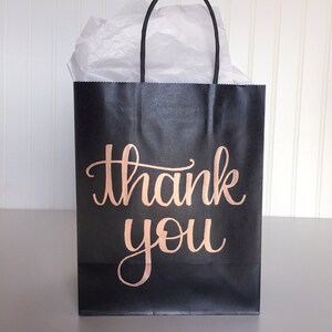 May include: Black paper gift bag with a rose gold "thank you" inscription. The bag is filled with white tissue paper.