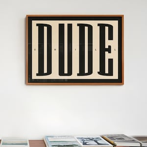 May include: A wooden shelf unit with multiple shelves filled with books. A framed print with the word "DUDE" in black letters on a white background hangs on the wall behind the shelf.