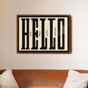 以下が含まれることがあります： 「HELLO」という単語が大きく黒い文字で書かれた額装されたプリント。文字は、「チャオ」、「オラ」、「アロハ」、「ボンジュール」など、さまざまな言語の小さな単語で構成されています。