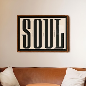 May include: A framed print with the word "SOUL" in large black letters. The letters are divided into four sections, each with a word: "RESPECT", "PASSION", "HARMONY", and "TRUST". The print is hanging on a light beige wall above a brown leather couch.