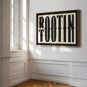 May include: A framed print with a black and white design of the word "ROOTIN' TOOTIN'" in a bold, stylized font. The print is framed in a light brown wood frame and is hanging on a white wall.