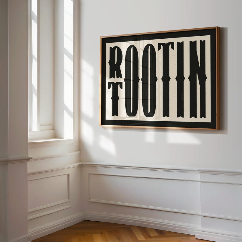 May include: A framed print with a black and white design of the word "ROOTIN' TOOTIN'" in a bold, stylized font. The print is framed in a light brown wood frame and is hanging on a white wall.