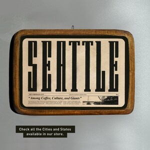 May include: A wooden sign with a vintage newspaper design featuring the word "SEATTLE" in bold black letters. The sign also includes the text "THE EMERALD CITY" and the quote "Among Coffee, Culture, and Giants".