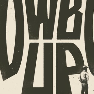 May include: Black and white graphic design with the text "COWBOY UP" in bold, block letters. A person wearing a cowboy hat and a long-sleeved shirt stands in front of the text.