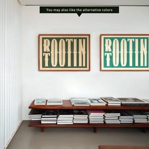May include: Two framed prints with the word "ROOTIN" in a stylized font. The prints are in different colors, one with a green background and the other with a blue background. The prints are hanging on a white wall above a wooden shelf with books.