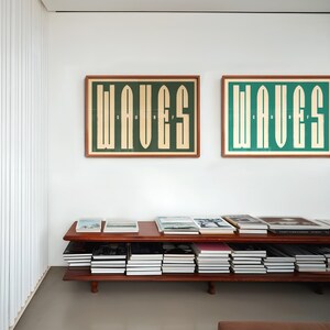 May include: Two framed prints with the word "WAVES" in a bold, graphic font. The prints are hung on a white wall above a wooden shelf filled with books.