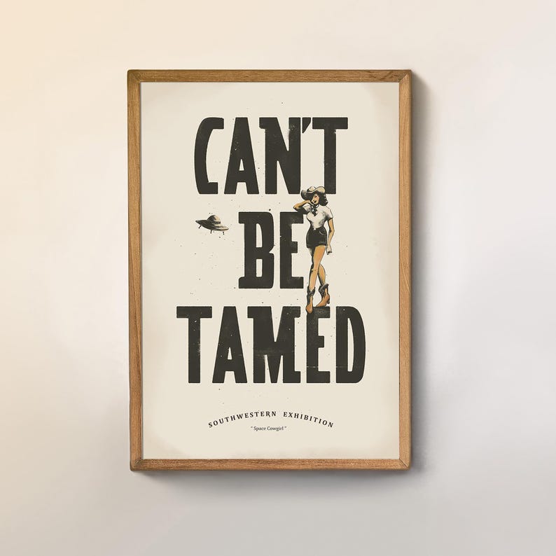 Pu&ograve; includere: Una stampa incorniciata con un design grafico in bianco e nero che dice "Can't Be Tamed" con una donna con un cappello da cowboy e un disco volante sullo sfondo. Il testo "Southwestern Exhibition" &egrave; sotto il design grafico.