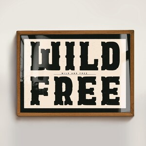 May include: A framed print with a black and white design. The text reads "WILD AND FREE" with the word "WILD" in large, bold letters and the word "FREE" in smaller, bold letters.