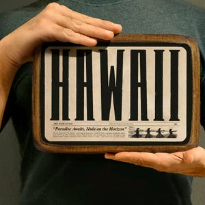 Op de afbeelding: Een houten bord met het woord "HAWAII" in grote zwarte letters. Het bord is versierd met een krantenontwerp en de tekst "The Aloha State "Paradise Awaits, Hula on the Horizon" en een illustratie van hula-dansers.