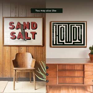 May include: Two framed prints on a wooden wall. The print on the left is a red and white graphic with the text "SAND SALT" and a surfboard. The print on the right is a black and white graphic with the text "HOLIDAY" in a maze pattern.