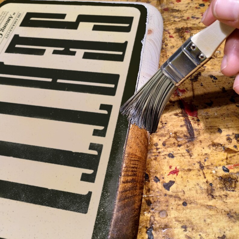 May include: A close-up of a wooden sign with a black stencil design. A person is using a brush to apply a dark brown stain to the edges of the sign.