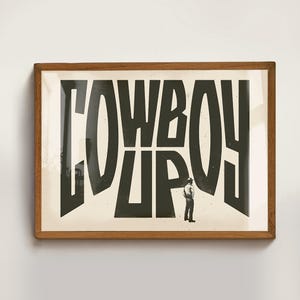 May include: A framed print with the text "COWBOY UP" in black lettering on a white background. A small figure of a person in a cowboy hat stands in front of the text.