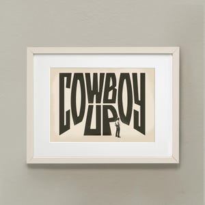 May include: A framed print with a black and white illustration of a cowboy standing in front of the word "COWBOY" written in large, stylized letters.