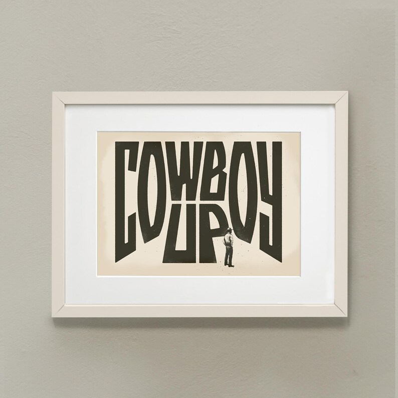 May include: A framed print with a black and white illustration of a cowboy standing in front of the word "COWBOY" written in large, stylized letters.