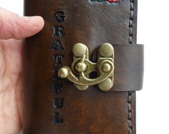 May include: A brown leather journal with a clasp closure and the word "GRATEFUL" embossed on the front. The journal is decorated with two red roses with green leaves.