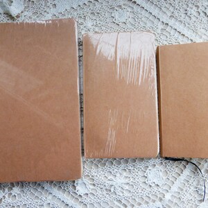 May include: Three brown notebooks of different sizes, wrapped in plastic, on a white lace tablecloth.