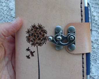 May include: A light tan leather journal with a dandelion design. The journal has a metal clasp closure and a pen holder. The dandelion is etched in brown, with a black stem and three flying seeds. The journal is approximately 5 inches tall and 3 inches wide.