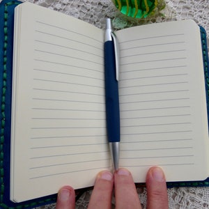 May include: A blue leather-bound journal with green stitching and a silver pen. The journal is open to a blank page with lined paper.