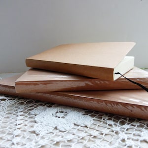 May include: Three brown notebooks stacked on top of each other. The top notebook has a black ribbon bookmark. The notebooks are on a white crocheted surface.