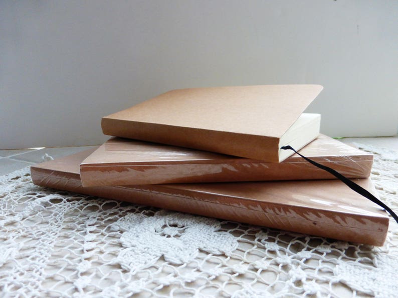 May include: Three brown notebooks stacked on top of each other. The top notebook has a black ribbon bookmark. The notebooks are on a white crocheted surface.