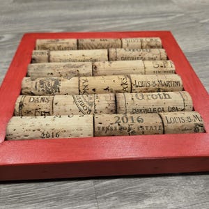 May include: A red wooden frame with a collection of wine corks arranged in rows. The corks are from various wineries and include the names "Dans", "Robert", "Silver Oak", "Groth", "Tesseon Estate", and "Louis M. Martini".