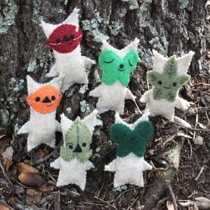 May include: A collection of handmade felt creatures with white bodies and various colored masks and leaf-like features. The creatures have orange, red, green, and leaf-shaped accents. They are arranged on a tree trunk.