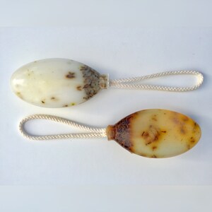 May include: Two oval-shaped soap bars with a string attached. The soap bars are translucent and have a light brown color with flecks of green and brown throughout. The string is white and is tied to the top of each soap bar.