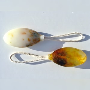 May include: Two oval-shaped soap bars with natural ingredients and a white cotton rope attached. The soap on the left is white with brown and green flecks. The soap on the right is yellow with brown flecks.