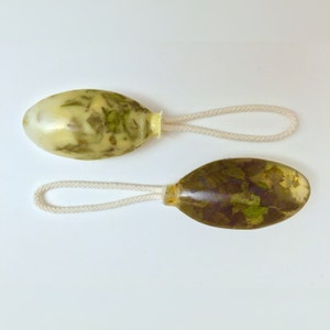 May include: Two oval-shaped soaps with a white string attached. The soaps are translucent and contain green and brown plant matter.
