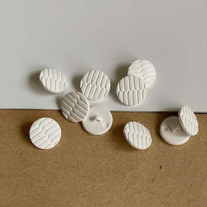 Peut inclure: Plusieurs boutons blancs et ronds avec un motif texturé et répétitif. Les boutons sont disposés sur une surface blanche et marron, mettant en valeur leur design et les légères variations de leur agencement. Les boutons ont un design classique et polyvalent.