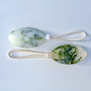 May include: Two oval-shaped bath soaps with green herbs suspended inside. The soaps are attached to white cotton cords.